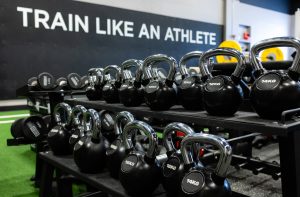 Kettlebells in a gym rack with weights labeled 8–16 kg, against a wall reading 'Train Like an Athlete' in the background