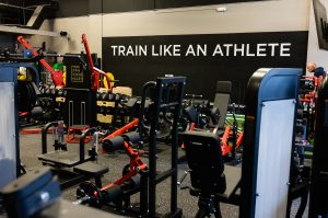 Gym interior filled with weight machines and red frames; a large wall reads 'TRAIN LIKE AN ATHLETE' in white letters.
