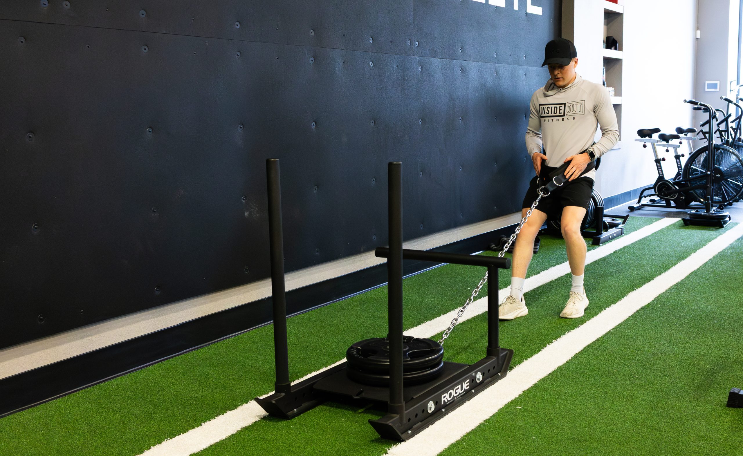 Man in a light gray Inside Out Fitness hoodie and cap performing a weighted sled drill on green turf in a gym.