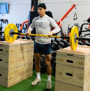 Man wearing a beanie lifting a barbell with yellow weight plates between wooden boxes in a gym. The Inside Out Fitness logo is visible on his white shirt.