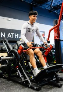 Young man in a light gray hoodie on a stationary bike, smiling in a gym. (informative)