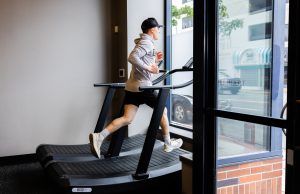 Man wearing a gray jacket and cap runs on a treadmill beside a large window in a gym.
