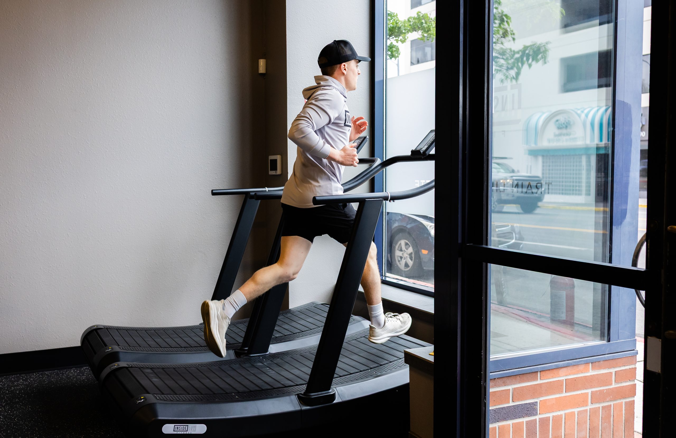 CM4A1232 | Inside Out Fitness Man wearing a gray jacket and cap runs on a treadmill beside a large window in a gym.