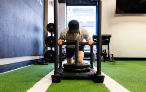 Person wearing a cap and gray hoodie performing a low push-up on a weight sled in a gym with green turf beneath.