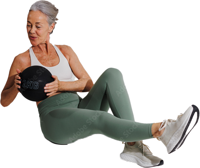 Senior woman in athletic wear sits on the floor, holding a small black medicine ball labeled 2KG, her legs bent in a workout pose with sneakers on.